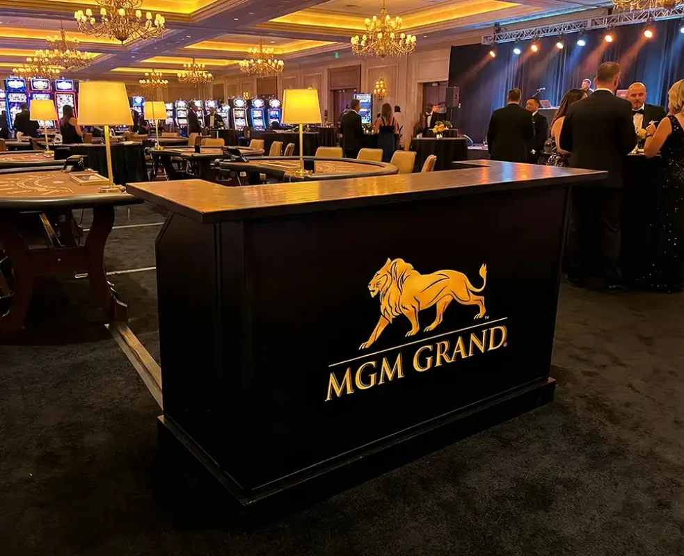カジノフロアのブラックジャックテーブルとイベント参加者の中に配置された、ゴールドのMGMグランドライオンロゴが光る黒いカスタムポータブルバーカウンター。.
